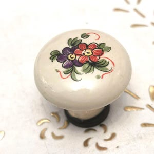 Pode incluir: Um puxador de armário de cerâmica creme com um design floral. O puxador apresenta um grupo de flores roxas e vermelhas com folhas verdes. A base é marrom escuro. Ferragens decorativas para móveis.