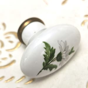 Peut inclure: Bouton de porte ovale en céramique blanche avec une base en métal doré et un motif de marguerite peint. La marguerite a des pétales blancs et un centre jaune, avec des feuilles vertes. Un accent décoratif pour les armoires ou les tiroirs.