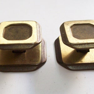 May include: Two brass cabinet knobs with a square shape and a raised octagonal centre. The knobs have a dark brown finish.