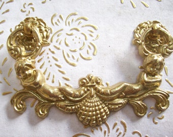 May include: Gold-toned metal drawer pull with a decorative design featuring two cherubs and a central floral motif. The pull has a ring handle.