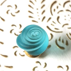 Puede incluir: Un pomo de armario de cristal en forma de rosa de color azul turquesa con base dorada.