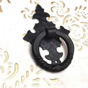 Op de afbeelding: Zwarte metalen ladeknop met een ringgreep. De decoratieve hardware heeft een sierlijke, bloemgeïnspireerde achterplaat. De ringgreep heeft een textuur, touwachtig ontwerp. De ladeknop staat tegen een witte achtergrond met gouden accenten.