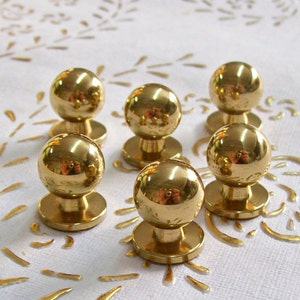 Könnte beinhalten: Sechs goldfarbene Metallknöpfe mit runder Oberseite und einer Basis. Die Knöpfe sind in einem Halbkreis auf einer weißen Oberfläche angeordnet.