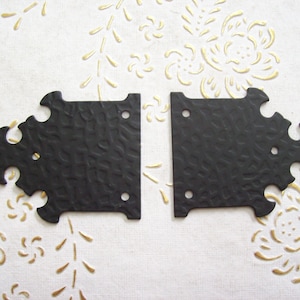 May include: Two black metal decorative corner brackets with a textured surface and ornate edges. Each bracket has three pre-drilled holes for mounting. Suitable for furniture or craft projects.