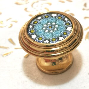 Puede incluir: Un pomo decorativo dorado con un diseño floral. La parte superior circular presenta un mosaico de patrones florales azules, amarillos y blancos. La base tiene forma redondeada y de pedestal.