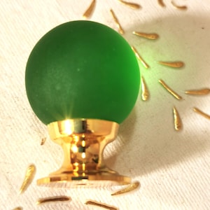 Op de afbeelding: Een groene glazen kastknop met een gouden basis. De knop is rond en heeft een gladde, matte afwerking.