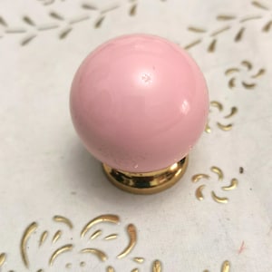 Puede incluir: Un pomo de armario esférico rosa con acabado brillante. El pomo está montado sobre una base dorada. El fondo es una superficie blanca con motivos florales dorados.