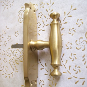 Może przedstawiać: Złoty klamka do drzwi z dźwignią. Klamka ma prostokątny element z dwoma otworami i zakrzywioną dźwignię z zaokrąglonym pokrętłem na końcu.