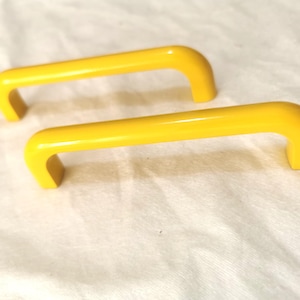 Può includere: Due maniglie per armadietti gialle brillanti e curve. Ogni maniglia ha una barra rettangolare arrotondata con estremità curve. Le maniglie sono realizzate in materiale lucido e sono su sfondo bianco.