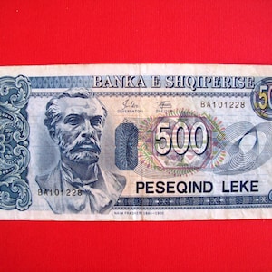 May include: A 500 Lek Albanian banknote featuring a portrait of Naim Frashëri, a prominent Albanian writer and poet. The banknote is blue, green, and red with a stylized design.