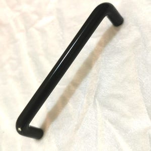Peut inclure: Une poignée de meuble noire avec une barre rectangulaire et des extrémités incurvées. La poignée est faite d'un matériau brillant et est posée sur un fond blanc.
