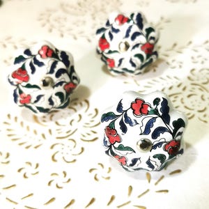 Peut inclure: Trois boutons en céramique blanche avec un motif floral dans les tons rouge, bleu et vert. Les boutons ont un bord festonné et une vis en métal argenté au centre.