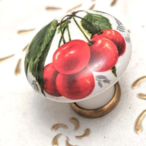 May include: A white ceramic cabinet knob with a design of red cherries and green leaves. The knob has a gold-coloured base and a white stem. Decorative hardware for furniture.