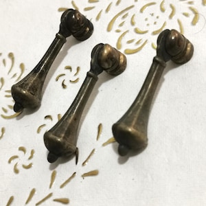 Op de afbeelding: Drie antieke messing kastknoppen met een afgeronde bovenkant en een lang, slank, taps toelopend lichaam. De knoppen zijn in een driehoekige vorm op een wit oppervlak gerangschikt.