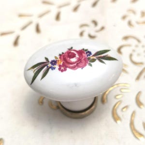 May include: White oval ceramic cabinet knob with a floral design featuring a large red rose and smaller purple and yellow flowers. The knob has a brass base, suitable for furniture or decor.