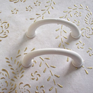 May include: Two white plastic handles with a rounded shape. The handles are on a white surface with a gold floral pattern.