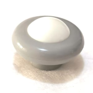 Pode incluir: Um puxador de armário cinza e branco em forma de cogumelo. O puxador tem um topo arredondado com um centro branco e um anel externo cinza. A base tem uma forma cilíndrica curta. O puxador é provavelmente feito de plástico ou material semelhante.