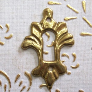 Peut inclure: Une plaque de trou de serrure ornée de couleur laiton avec un motif floral décoratif. L'écusson a la forme d'un trou de serrure avec un trou en haut et en bas. Le fond est une surface blanche texturée avec des embellissements dorés.