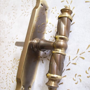 May include: A brass door handle with a decorative twisted design. The handle has a rectangular plate with rounded edges and a T-shaped lever.