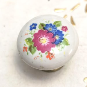 Puede incluir: Pomo de armario de cerámica blanca con diseño floral. Las flores son rosas, azules y naranjas con hojas verdes. El pomo tiene una base de latón, para cajón o armario.