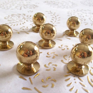 Könnte beinhalten: Sieben runde, goldfarbene Schrankknöpfe mit glatter, glänzender Oberfläche. Die Knöpfe sind in einem Halbkreis auf einer weißen Oberfläche angeordnet.