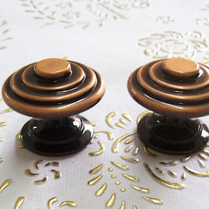 May include: Two round, copper-toned cabinet knobs with a black base. Each knob has four concentric rings.