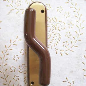 May include: A brown door handle with a gold-coloured backplate. The handle has a curved shape and is mounted on a white surface with a floral pattern.