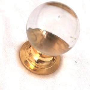 Può includere: Un pomello per porta in vetro trasparente con base dorata e semisfera dorata. Il pomello è rotondo e ha una finitura lucida. La base è circolare e ha una superficie liscia. Questo hardware decorativo è adatto per porte e armadi.