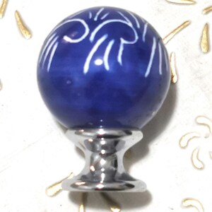 Op de afbeelding: Een kobaltblauwe keramische kastknop met een wit decoratief patroon. De knop heeft een bolvorm en is gemonteerd op een zilverkleurige metalen basis. Decoratieve accenten omringen de knop.