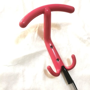 Op de afbeelding: Een felroze, T-vormige handgreep met twee gebogen haken en een doorzichtige plastic buis. De handgreep heeft een gebogen bovenkant en een gebogen steel met twee gaten. De haken bevinden zich aan de onderkant.