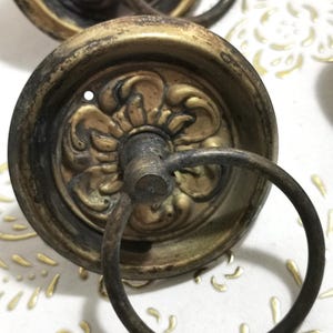 Pode incluir: Um par de puxadores de porta de armário de latão antigo com desenhos florais ornamentados e uma alça de anel. Os puxadores têm um acabamento desgastado e envelhecido.