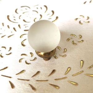 Può includere: Un pomello in vetro bianco con una base in metallo dorato. Il pomello è rotondo e ha una finitura opaca.