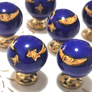 May include: A collection of blue ceramic cabinet knobs with gold accents. Each knob features a celestial design with gold stars and crescent moons. The knobs have a round shape and sit on a gold base.