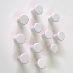May include: A set of 13 white ceramic knobs, each with a rounded top and a short stem. The knobs are arranged in a circular pattern on a white background.