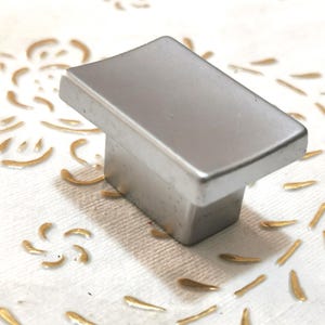 Puede incluir: Un pomo de armario de plata con forma rectangular y bordes redondeados.