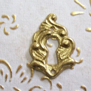 Può includere: Una copertura per buco della serratura dorata e ornata con un design decorativo a spirale. La copertura per buco della serratura è centrata su una superficie strutturata bianco sporco con abbellimenti dorati. La copertura per buco della serratura ha un'estetica vintage.