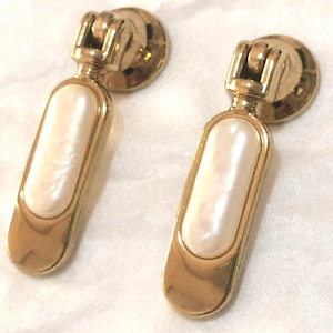 May include: Two gold-toned cabinet pulls with a rectangular, off-white, pearlescent center. The pulls have a circular base and a decorative top. The hardware is designed for furniture and adds a touch of elegance.