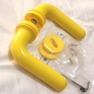 Op de afbeelding: Heldergele deurgrepen met bijpassende ronde afwerking en hardware. De set bevat twee handgrepen, verschillende afwerkingsstukken, een klein zakje met schroeven en een inbussleutel. De handgrepen zijn gebogen en hebben een gladde, glanzende afwerking.