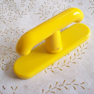 Puede incluir: Un mango de plástico amarillo brillante con una parte superior redondeada y una base rectangular. El mango está unido a la base con un soporte cilíndrico corto. Es probable que el objeto sea una herramienta o parte de un artículo más grande.