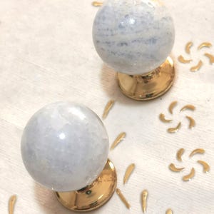 Puede incluir: Dos tiradores decorativos para armarios. Cada tirador presenta una piedra esférica azul claro sobre una base dorada. Los tiradores se colocan sobre un fondo blanco con detalles florales dorados, creando una estética elegante.