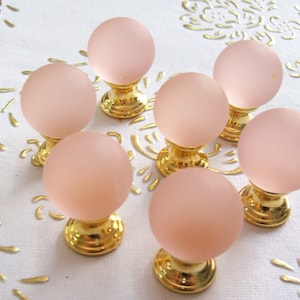 May include: Set of eight pink glass cabinet knobs with gold metal bases. The knobs are round and have a frosted finish.
