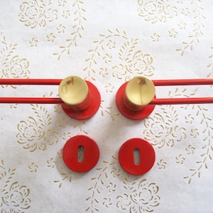 Może przedstawiać: Dwie czerwone klamki drzwiowe ze złotymi gałkami i dwie pasujące okrągłe czerwone rozety.