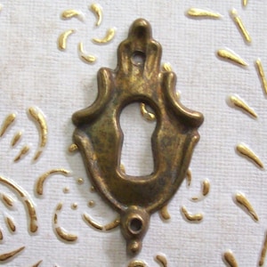 Può includere: Un copri-chiave in ottone con un design elaborato. Il copri-chiave è di colore oro chiaro e ha un buco della serratura al centro.