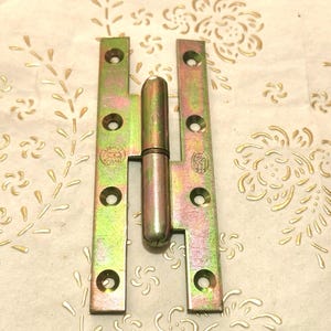 Pode incluir: Uma dobradiça de metal com um centro cilíndrico e duas placas retangulares. As placas são douradas e verdes com vários orifícios para parafusos. A dobradiça é projetada para uso em móveis ou portas.