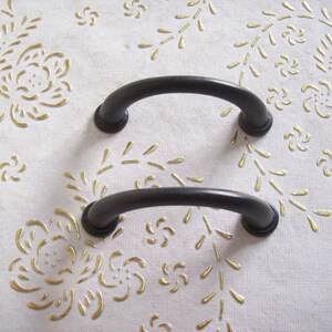 Pode incluir: Duas puxadores de porta de armário de metal preto com um design arqueado simples. As puxadores estão colocadas sobre um fundo branco com um padrão floral dourado.