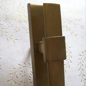 May include: A gold-toned door handle with a rectangular design. The handle has a square-shaped lever and a rectangular backplate.