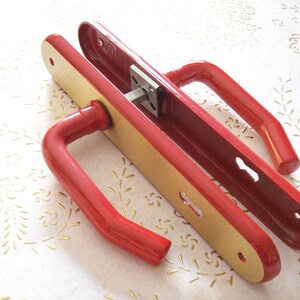 May include: Red door handle set with a gold-coloured plate. The handle is made of metal and has a simple, modern design.