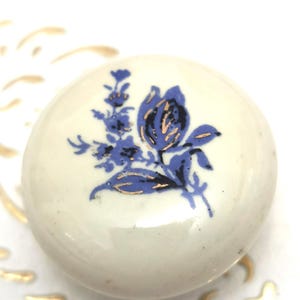 May include: A round, cream-coloured ceramic knob featuring a blue floral design with gold detailing. The floral pattern includes a large flower and smaller sprigs. The knob is likely for a cabinet or drawer and has a smooth, glossy finish.