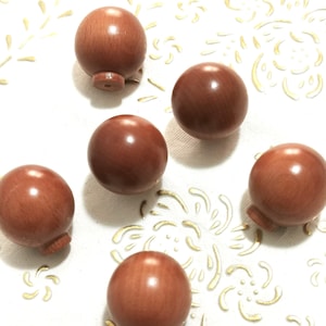Op de afbeelding: Zes ronde, gladde houten knoppen in roodbruine kleur. De knoppen zijn in een halve cirkel op een wit oppervlak met een gouden bloemenpatroon gerangschikt.