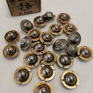 Puede incluir: Un conjunto de unidades de transferencia de bolas con base dorada y una bola de acero plateado. Están dispersas sobre una superficie clara. Una caja de cartón con la inscripción "Art. 5008 25 mm" es visible. El diámetro es de unos 2,5 cm.